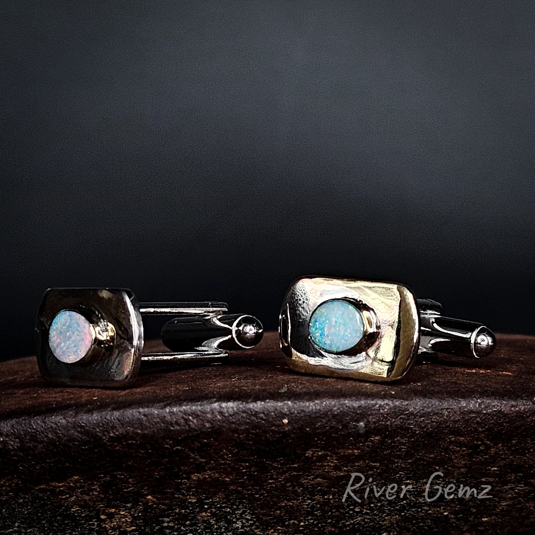 A pair of men's cufflinks by River Gemz, featuring textured square bezel settings in silver with light blue opal centers, displayed on a dark wooden surface against a dark background.