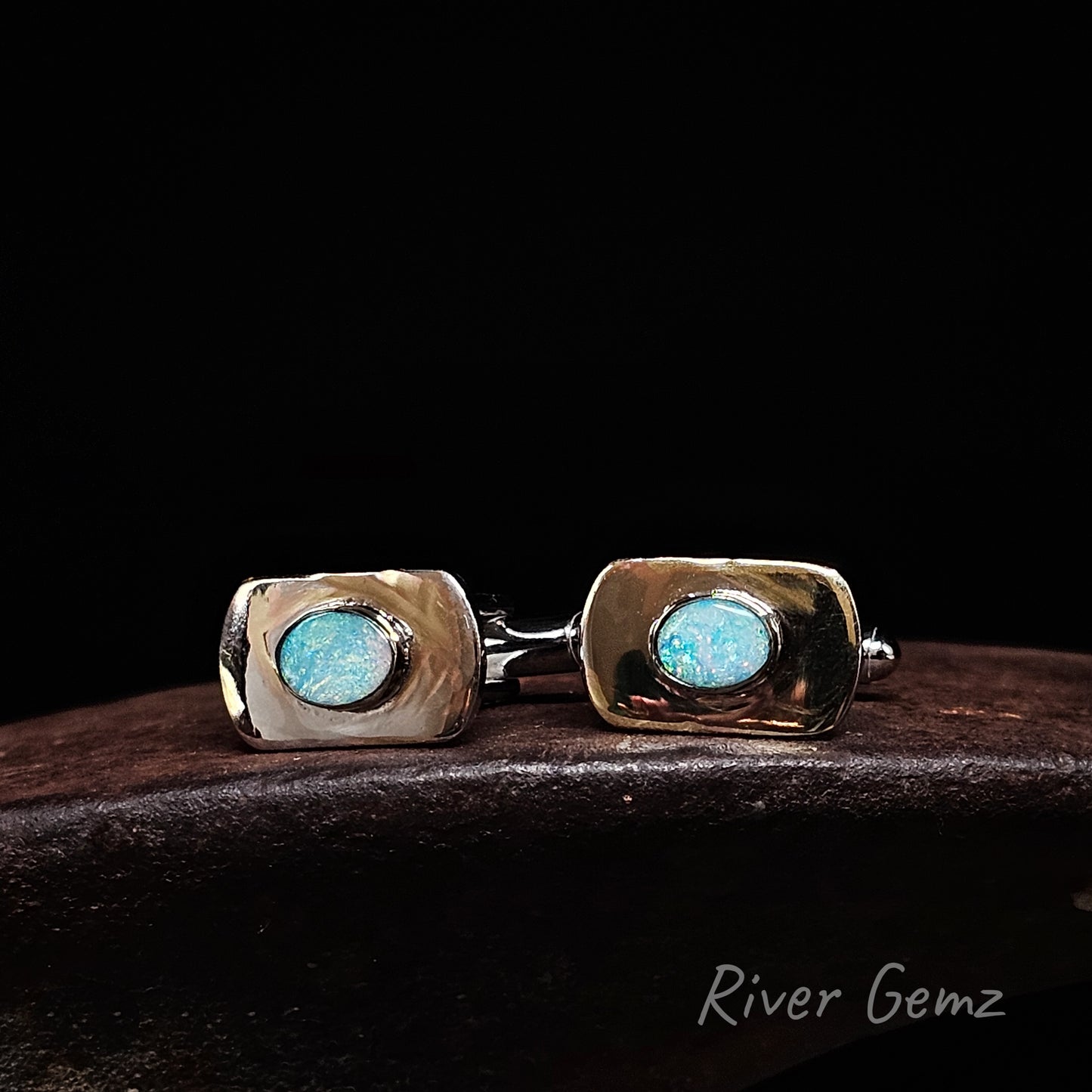 A pair of men's cufflinks with rectangular silver bezel settings holding round light blue gemstones, displayed on a dark wooden surface against a black background. The brand name 'River Gemz' is visible in the bottom right corner.