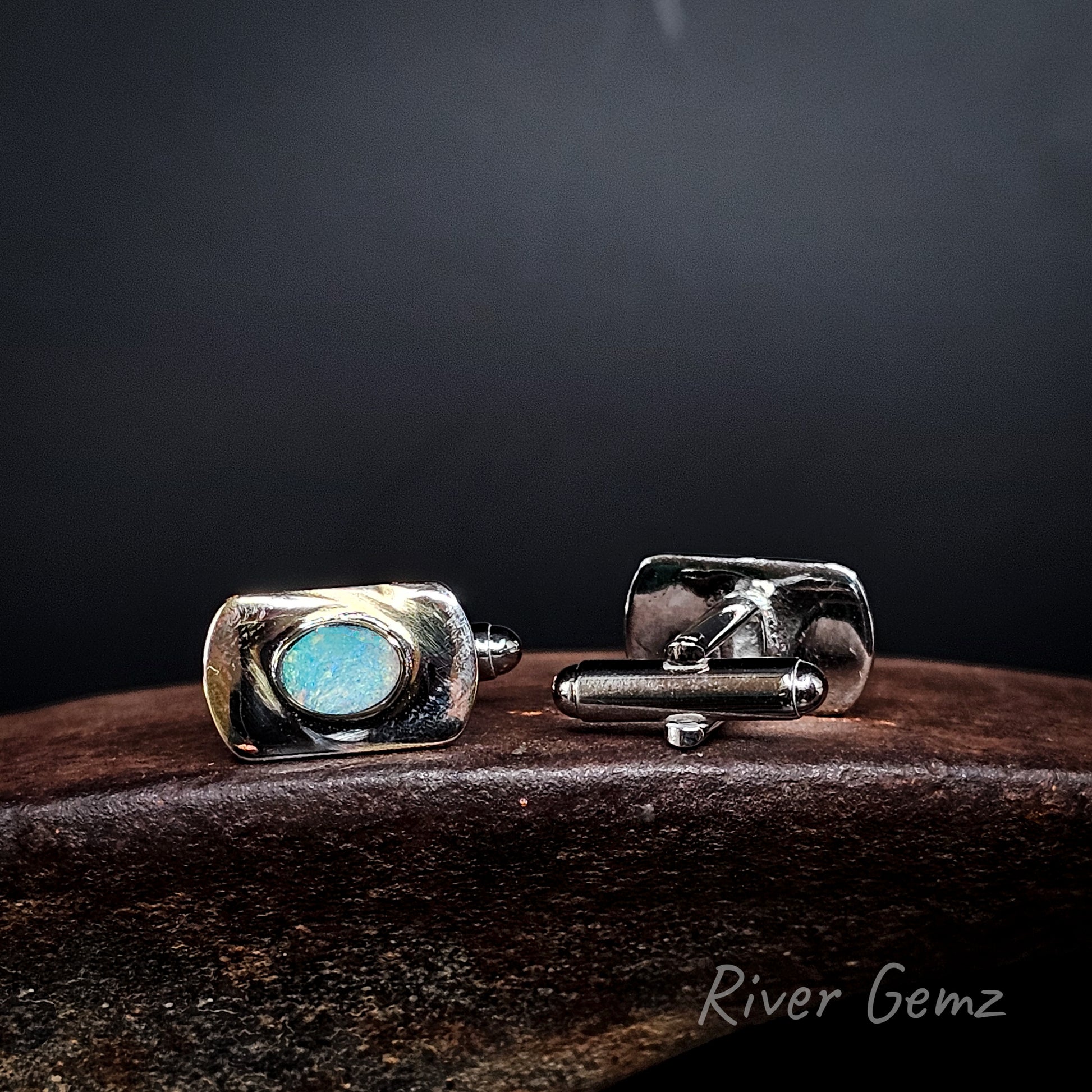 A pair of men's cufflinks by River Gemz, featuring a polished silver-tone metal body with a central oval light blue stone, displayed on a dark wooden surface against a dark background.