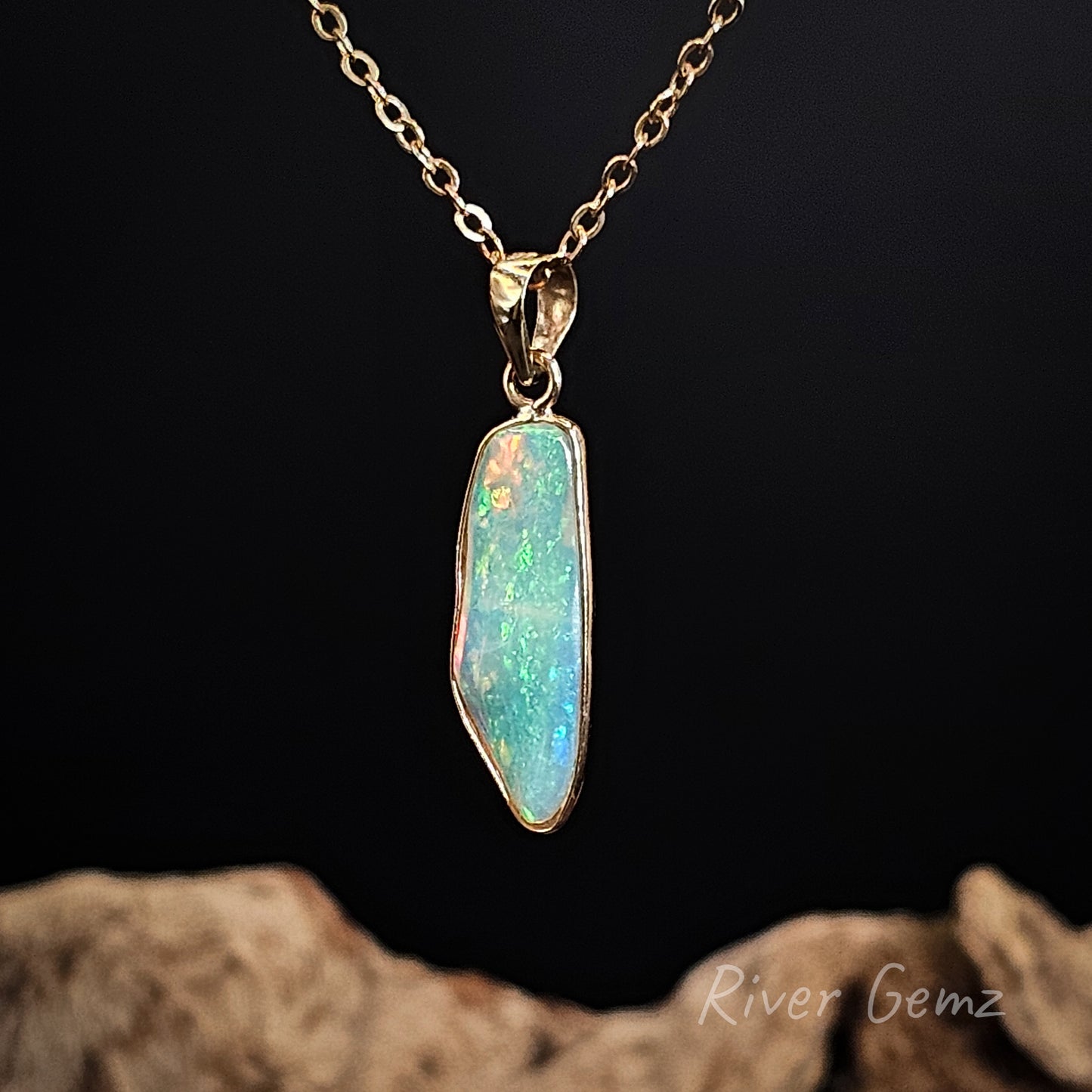 Light opal besel set in yellow gold pendant on a gold chain against a dark background with 'River Gemz' branding.