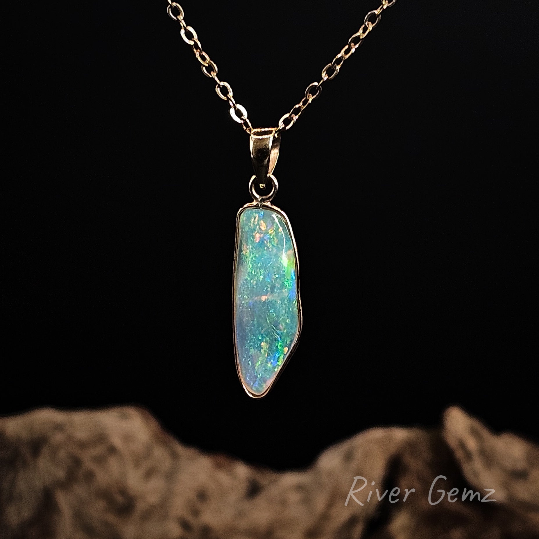 Multi-coloured shell opal besel set in yellow gold necklace on a dark background with 'River Gemz' branding.