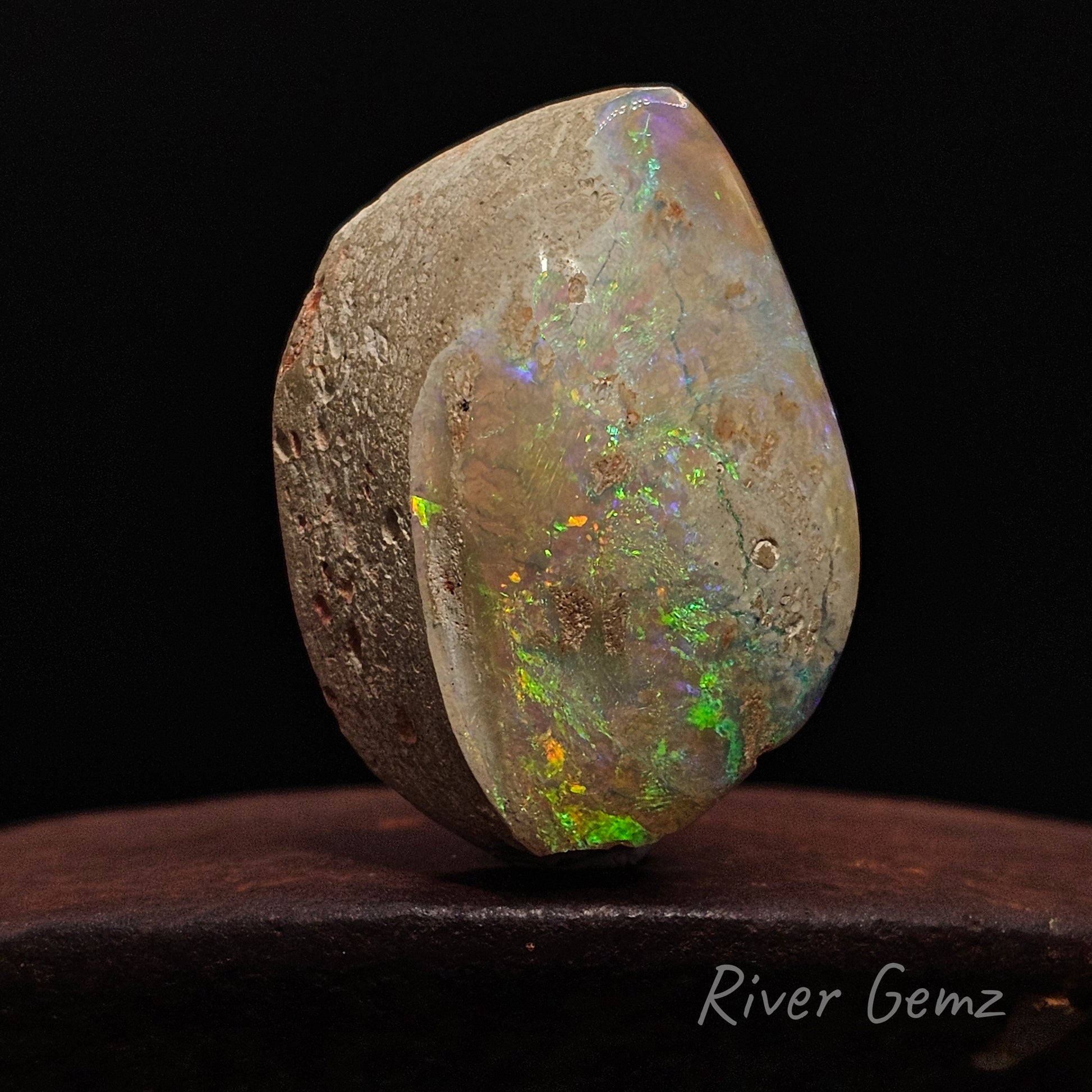 Opal stone with multicoloured play of light on a dark background, branded 'River Gemz'.