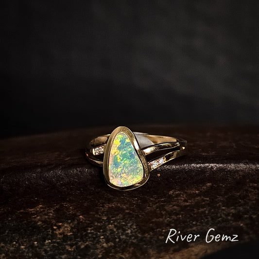 Gold ring with a free-form cut crystal opal stone and diamonds on a dark background, branded 'River Gemz'.