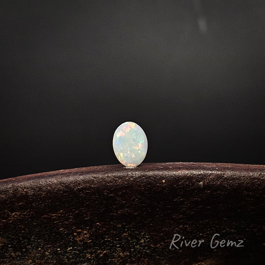 Oval shaped multi-coloured light opal standing upright on a weathered shoe anvil with 'River Gemz' watermark in the bottom right hand corner.