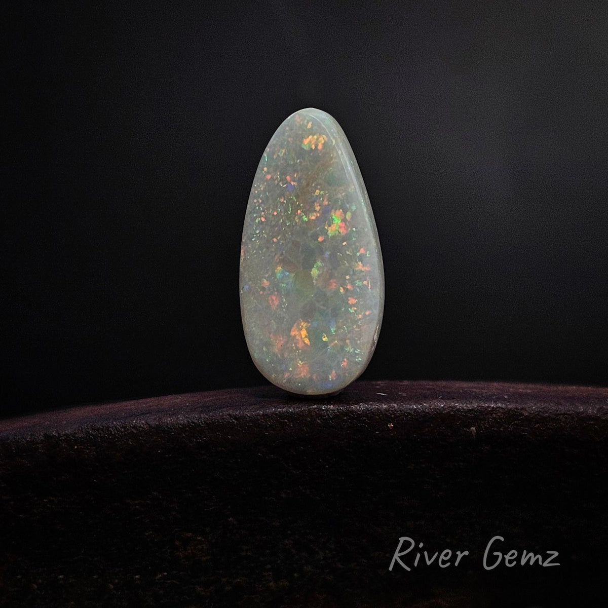 A teardrop-shaped white opal cabochon with vibrant multicolored flashes, displayed on a dark wooden surface against a black background. The brand name 'River Gemz' is visible in the bottom right corner.