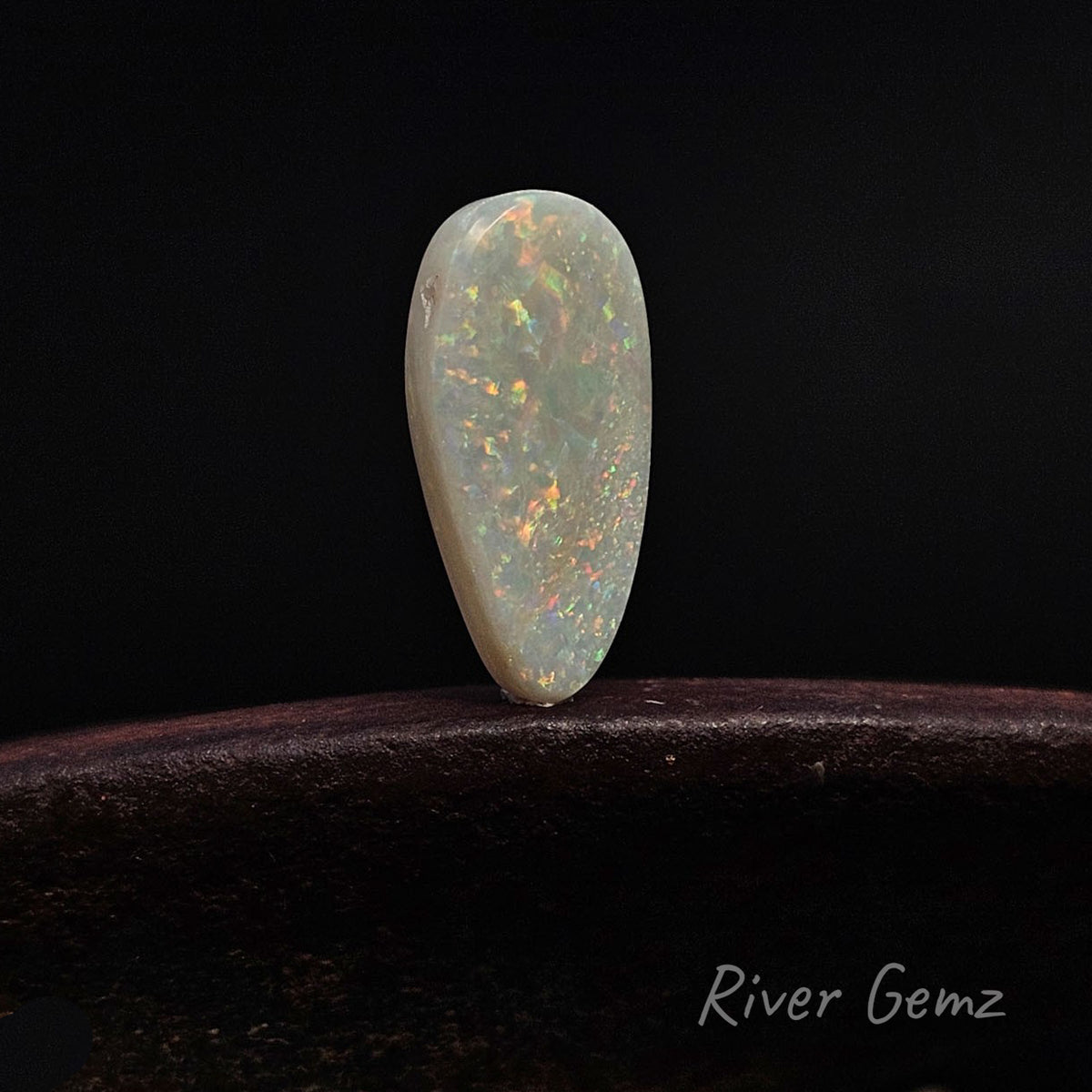 A teardrop-shaped white opal gemstone with vibrant internal flashes of orange, green, and blue, displayed on a dark wooden surface against a solid black background. The brand name 'River Gemz' is visible in the bottom right corner.