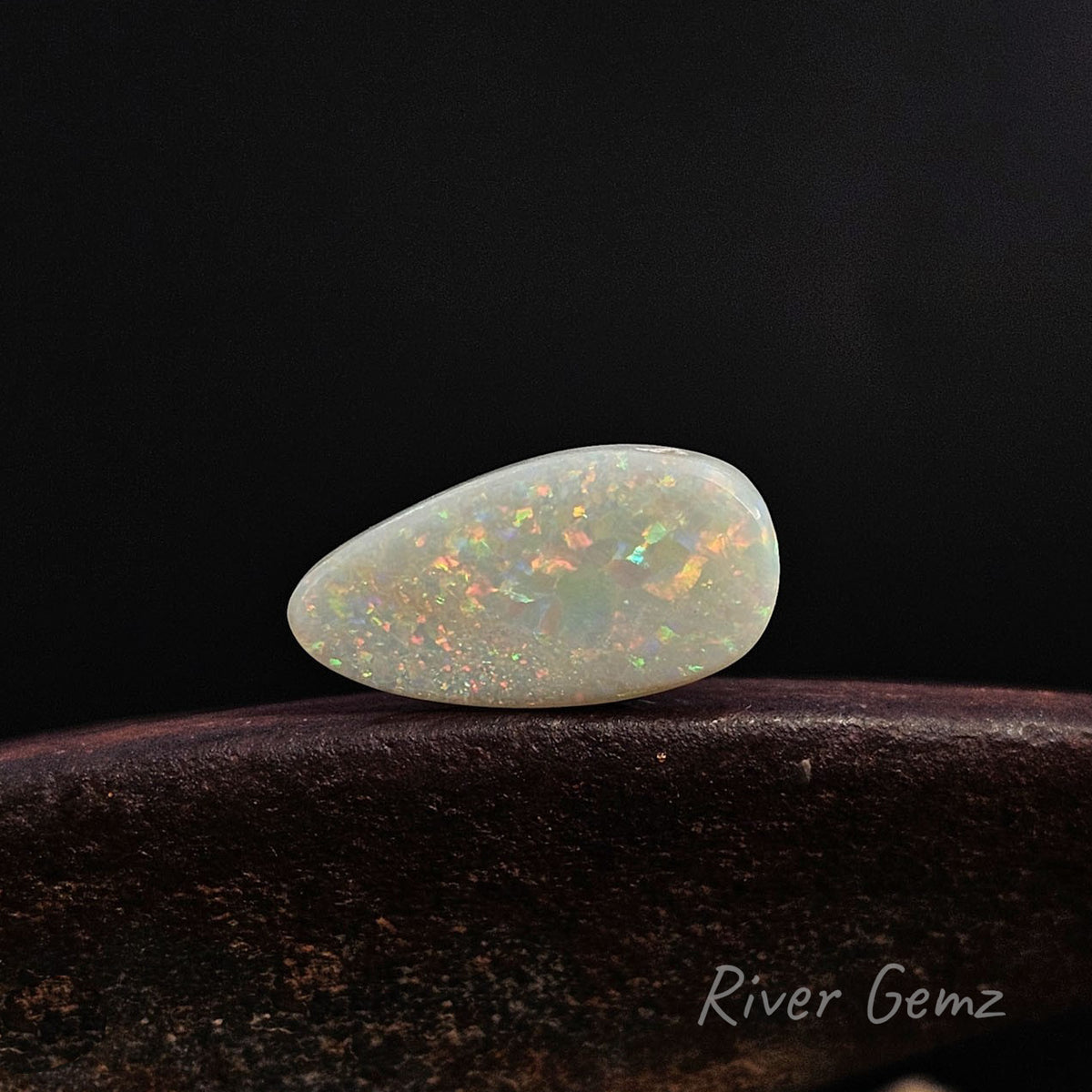 A polished teardrop-shaped white opal gemstone with vibrant iridescent flashes of orange, green, and blue, resting on a dark brown textured surface against a solid black background. The 'River Gemz' brand logo is visible in the bottom right corner.