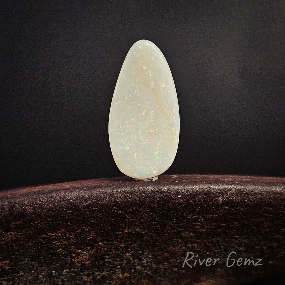 Rear view of a teardrop-shaped white opal gemstone showing the light base tone, standing upright on a dark wooden surface against a dark background. The 'River Gemz' logo is visible in the bottom right corner.