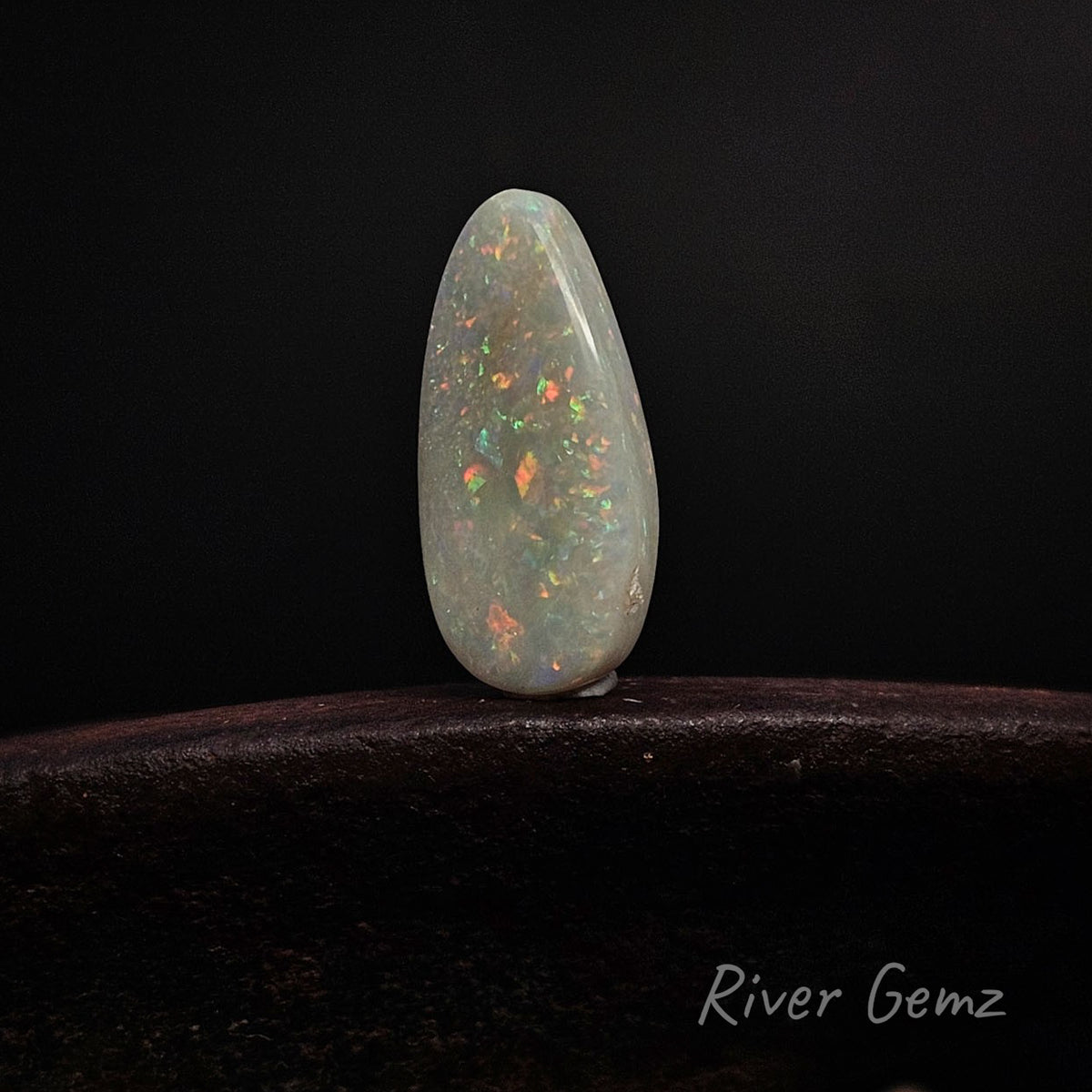 A teardrop-shaped white opal gemstone with vibrant orange, green, and blue flashes, displayed on a dark wooden surface against a black background. The brand name 'River Gemz' is visible in the bottom right corner.