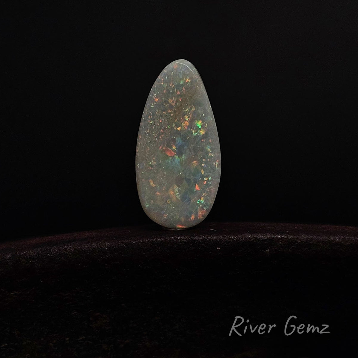A teardrop-shaped natural opal gemstone with a white base and vibrant multicolor flashes, standing upright on a dark surface against a black background. The brand name 'River Gemz' is visible in the bottom right corner.