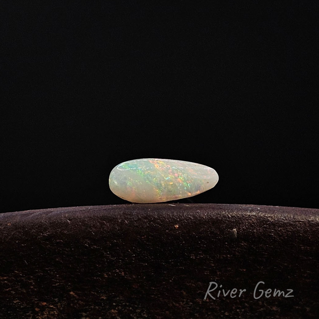Multi-coloured tear-drop shaped white opal lying horizontally on a brown surface.