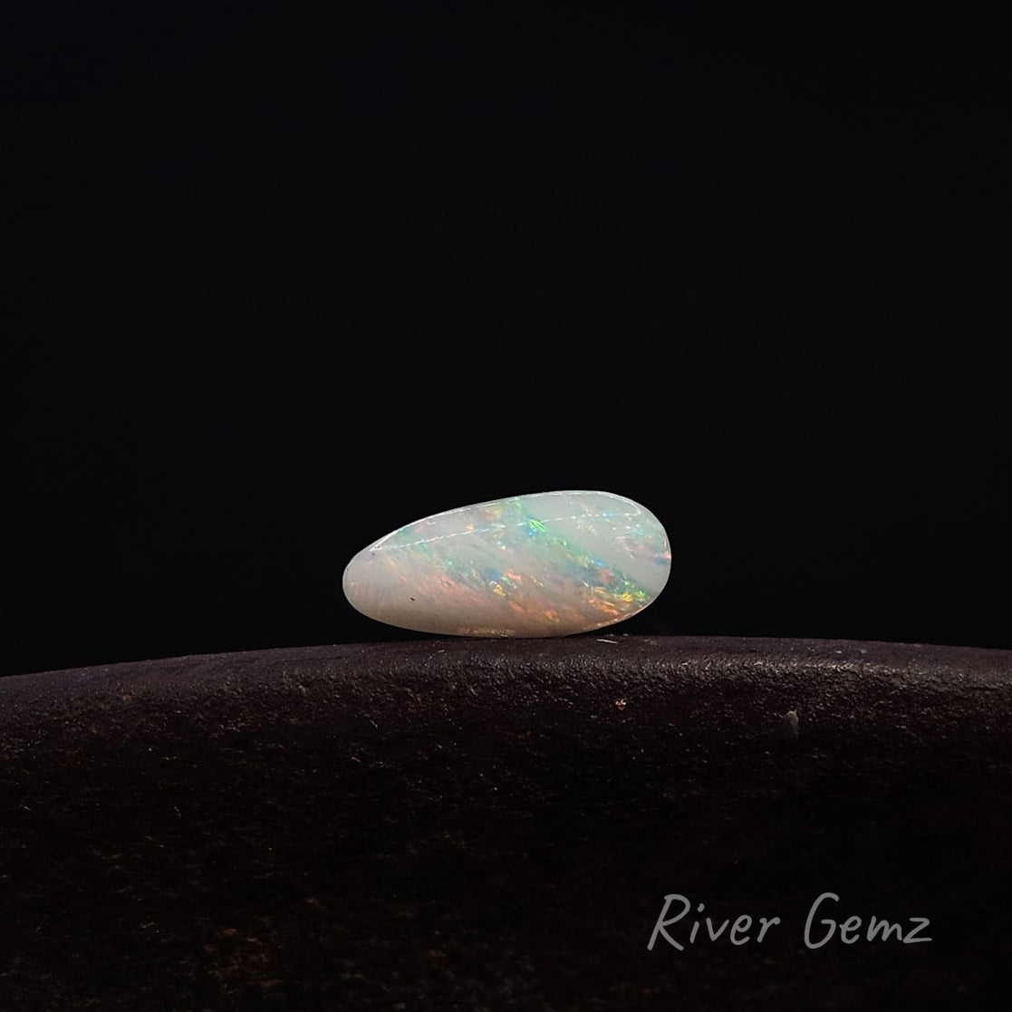 A polished white opal gemstone with vibrant multicolor play-of-color, resting on a dark textured surface against a solid black background. The brand name 'River Gemz' is visible in the bottom right corner.