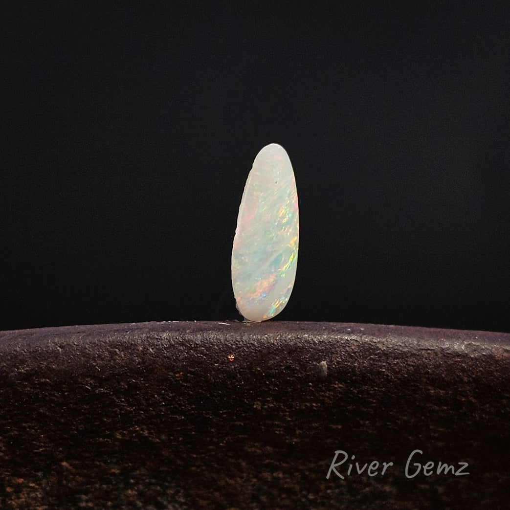 Rear view of a single polished oval opal gemstone with iridescent white, blue, and orange flashes, standing upright on a dark textured surface against a black background. The watermark 'River Gemz' is visible in the bottom right corner.