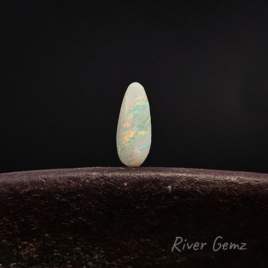 Tear drop shaped light opal with good variation in colour standing upright on weathered shoe anvil and 'River Gemz' watermark in the bottom right hand corner.