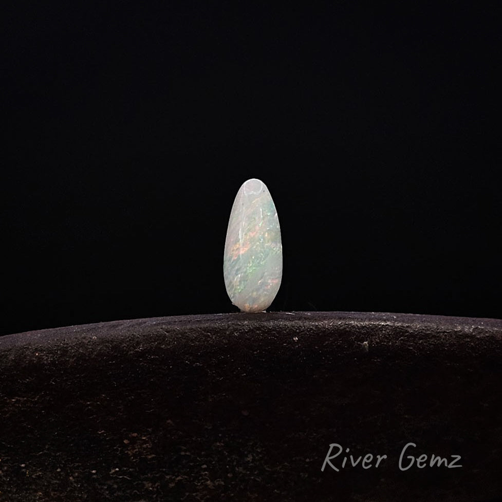 A polished teardrop-shaped opal gemstone with iridescent flashes of green, pink, and blue, standing upright on a dark textured surface against a solid black background. The 'River Gemz' brand logo is visible in the bottom right corner.