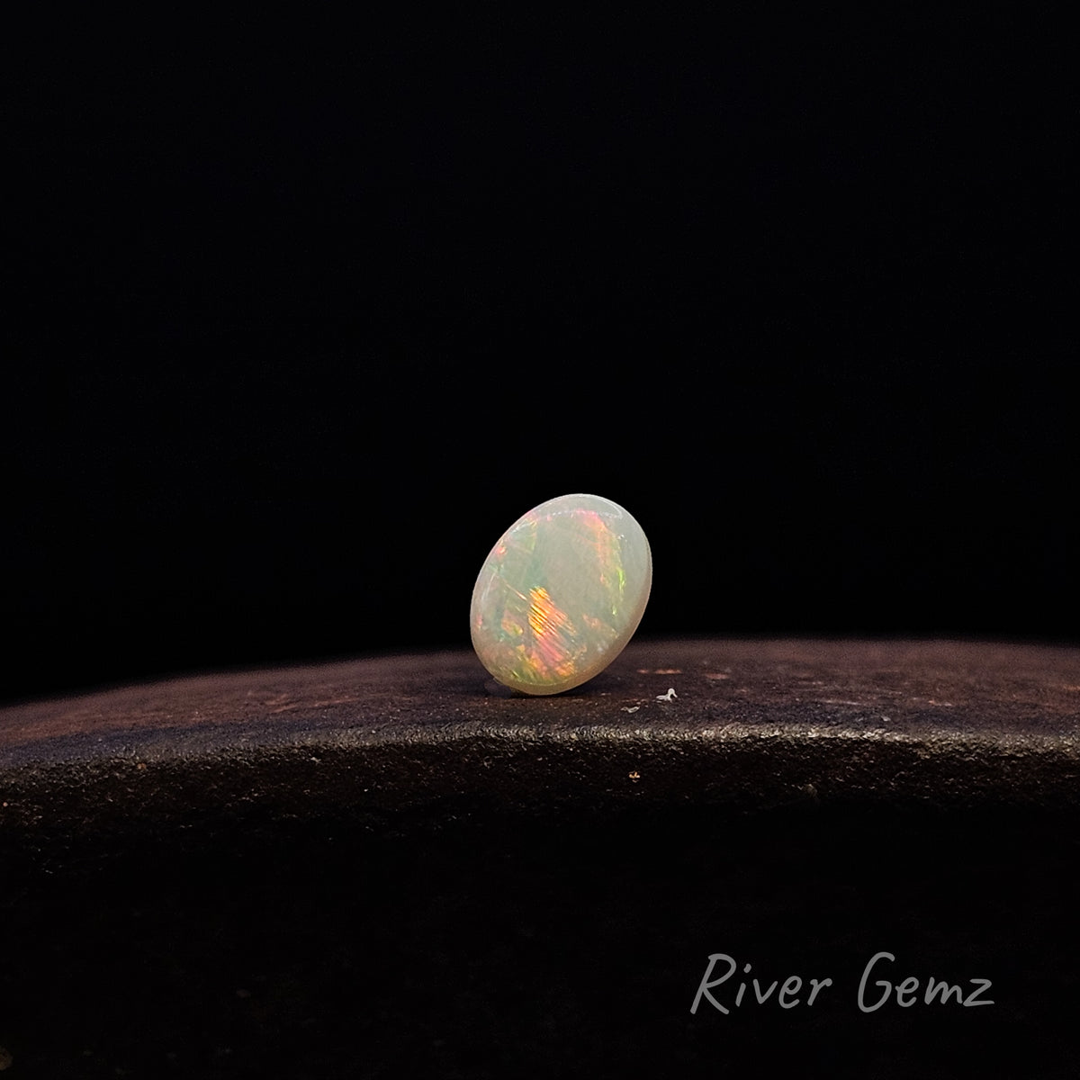 A small oval cabochon gemstone with iridescent opal-like play-of-color, standing on a dark textured surface against a black background. The River Gemz logo is visible in the bottom right corner.