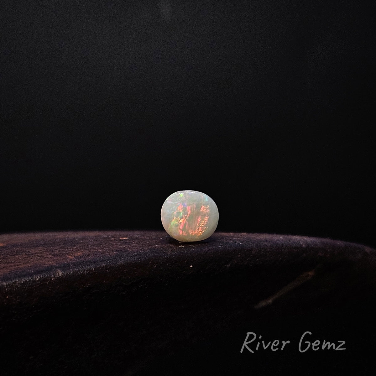 Oval shaped light opal positioned side ways on a dark surface. 'River Gemz' water mark in the bottom right hand corner.