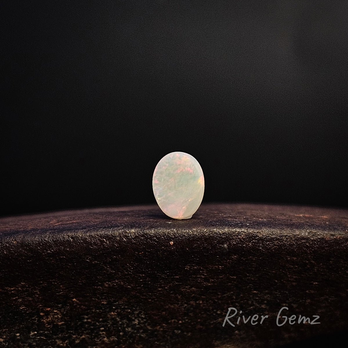 Rear view of a single oval cabochon gemstone with a milky white body and soft iridescent pink-blue flashes, standing upright on a dark textured surface against a black background. The watermark 'River Gemz' is visible in the bottom right corner.
