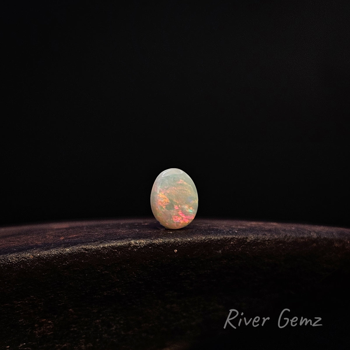 A small oval cabochon gemstone with a play-of-color effect, displaying white, peach, and orange hues, resting on a dark textured surface against a solid black background. The 'River Gemz' brand name is visible in the bottom right corner.