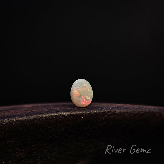 A small oval cabochon gemstone with a play-of-color effect, displaying white, peach, and orange hues, resting on a dark textured surface against a solid black background. The 'River Gemz' brand name is visible in the bottom right corner.