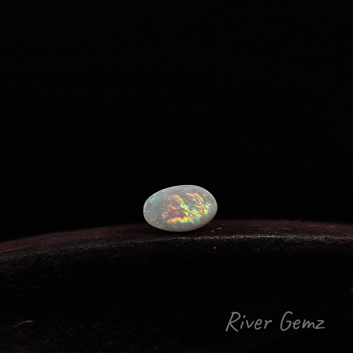 Oval shaped multi-coloured unset light opal shown lying E-W on dark brown surface with 'River Gemz' water mark shown in bottom right hand corner.