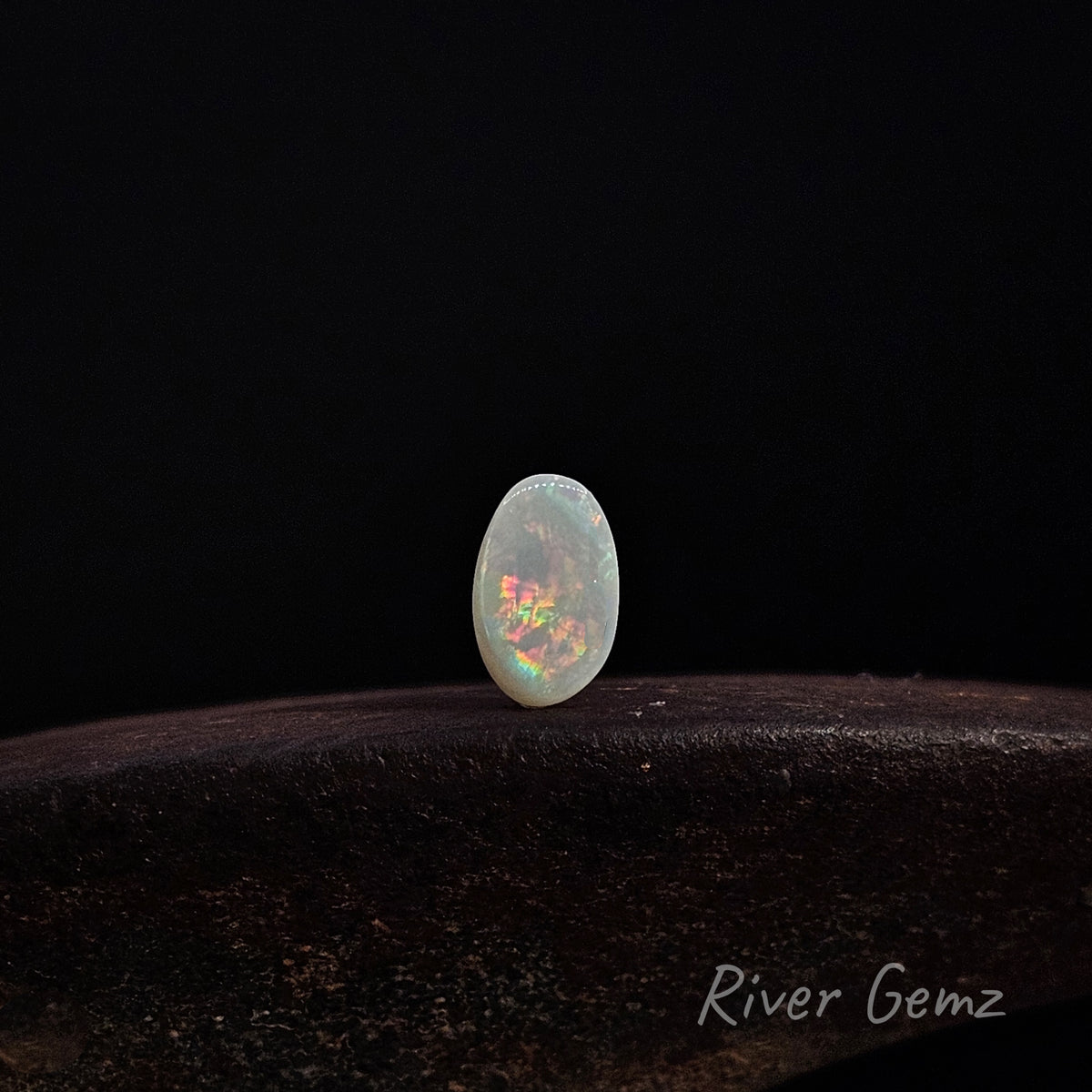 Oval shaped multi-coloured unset light opal standing upright on dark brown surface with 'River Gemz' water mark shown in bottom right hand corner.