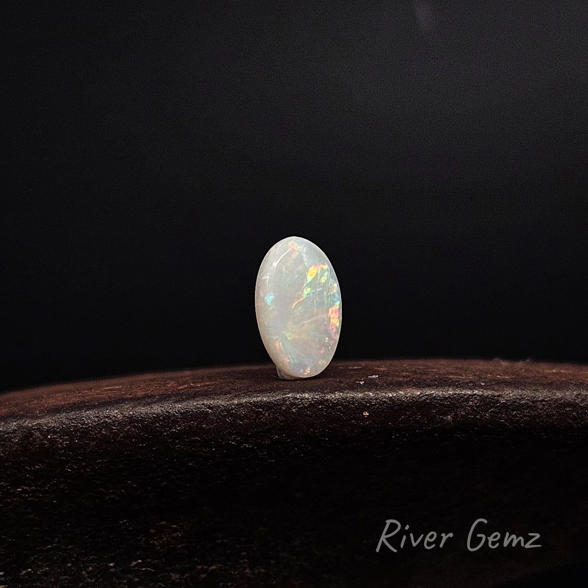 Oval shaped multi-coloured unset light opal standing upright on dark brown surface with 'River Gemz' water mark shown in bottom right hand corner.