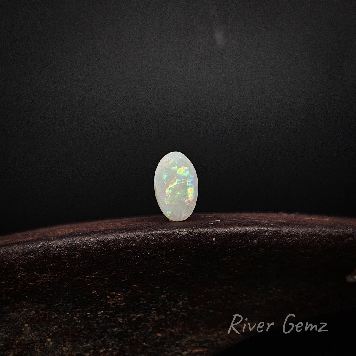 Oval shaped multi-coloured unset light opal standing upright on dark brown surface with 'River Gemz' water mark shown in bottom right hand corner.