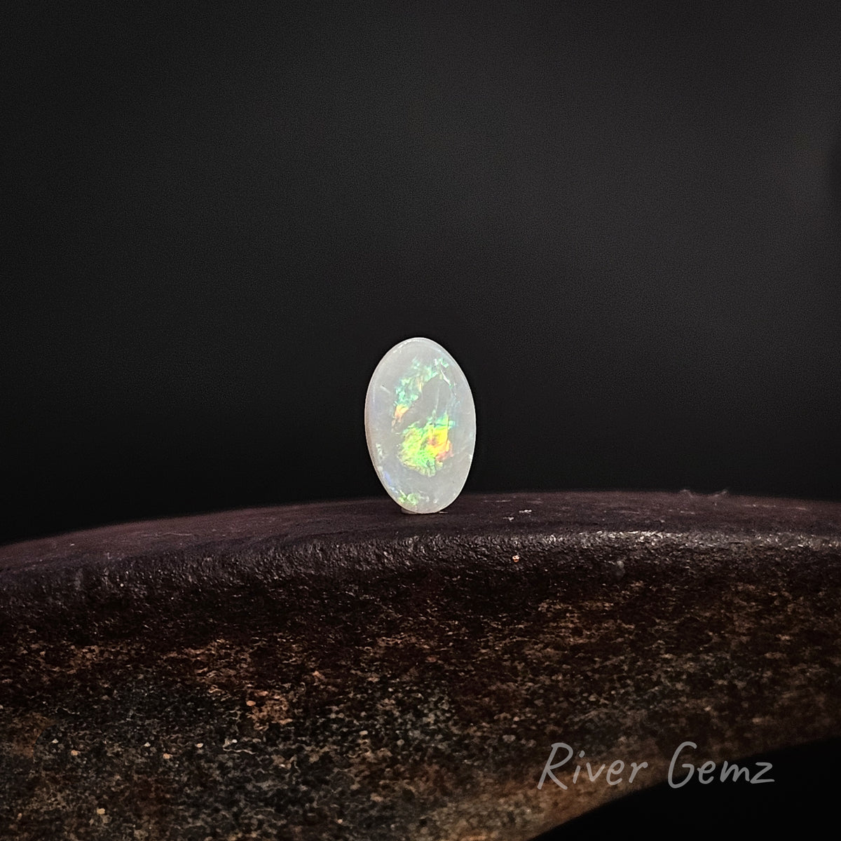 Oval shaped multi-coloured unset light opal standing upright on dark brown surface with 'River Gemz' water mark shown in bottom right hand corner.