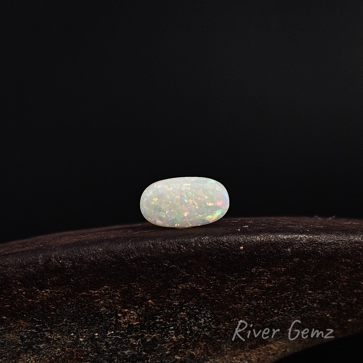 Oval shaped light opal in an East-West orientation on a brown surface with 'River Gemz' water mark in the bottom right hand corner.
