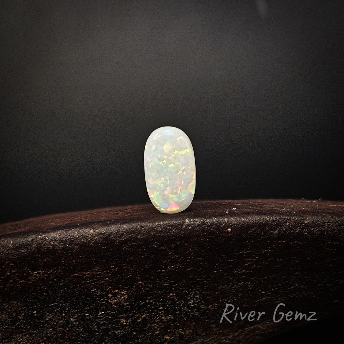 A small oval cabochon gemstone with a milky white body and vivid iridescent flashes of green, orange, and pink, displayed on a dark wooden surface against a dark gray background. The brand name 'River Gemz' is visible in the bottom right corner.