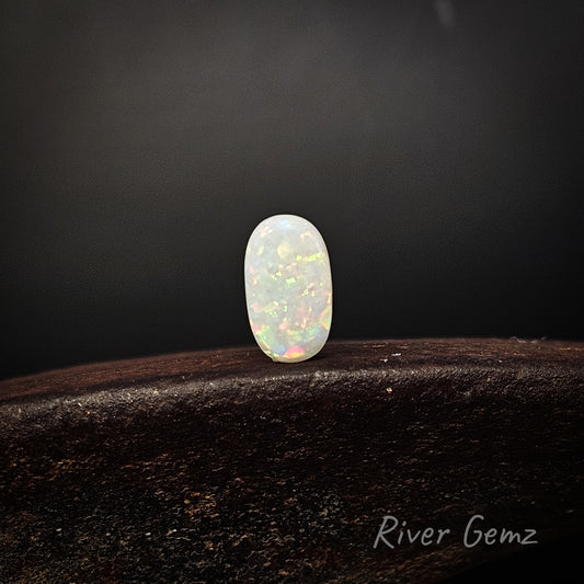 A small oval cabochon gemstone with a milky white body and vivid iridescent flashes of green, orange, and pink, displayed on a dark wooden surface against a dark gray background. The brand name 'River Gemz' is visible in the bottom right corner.