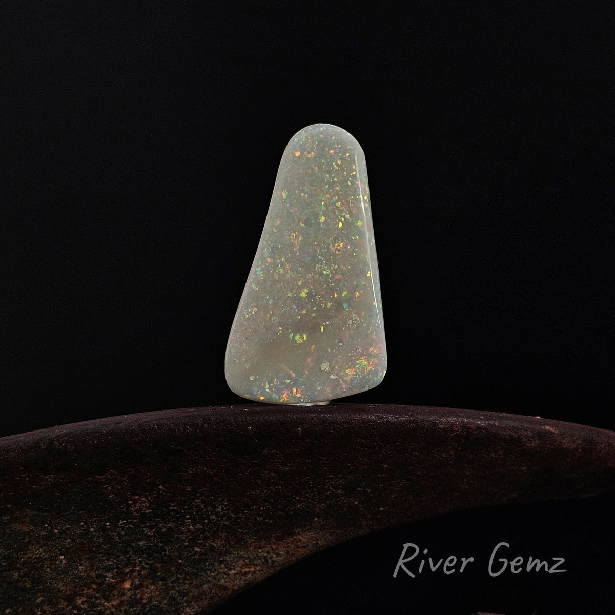 Multi-coloured free-form cut light opal on a shoe anvil against a black background and 'River Gemz' watermark in the bottom right hand corner.