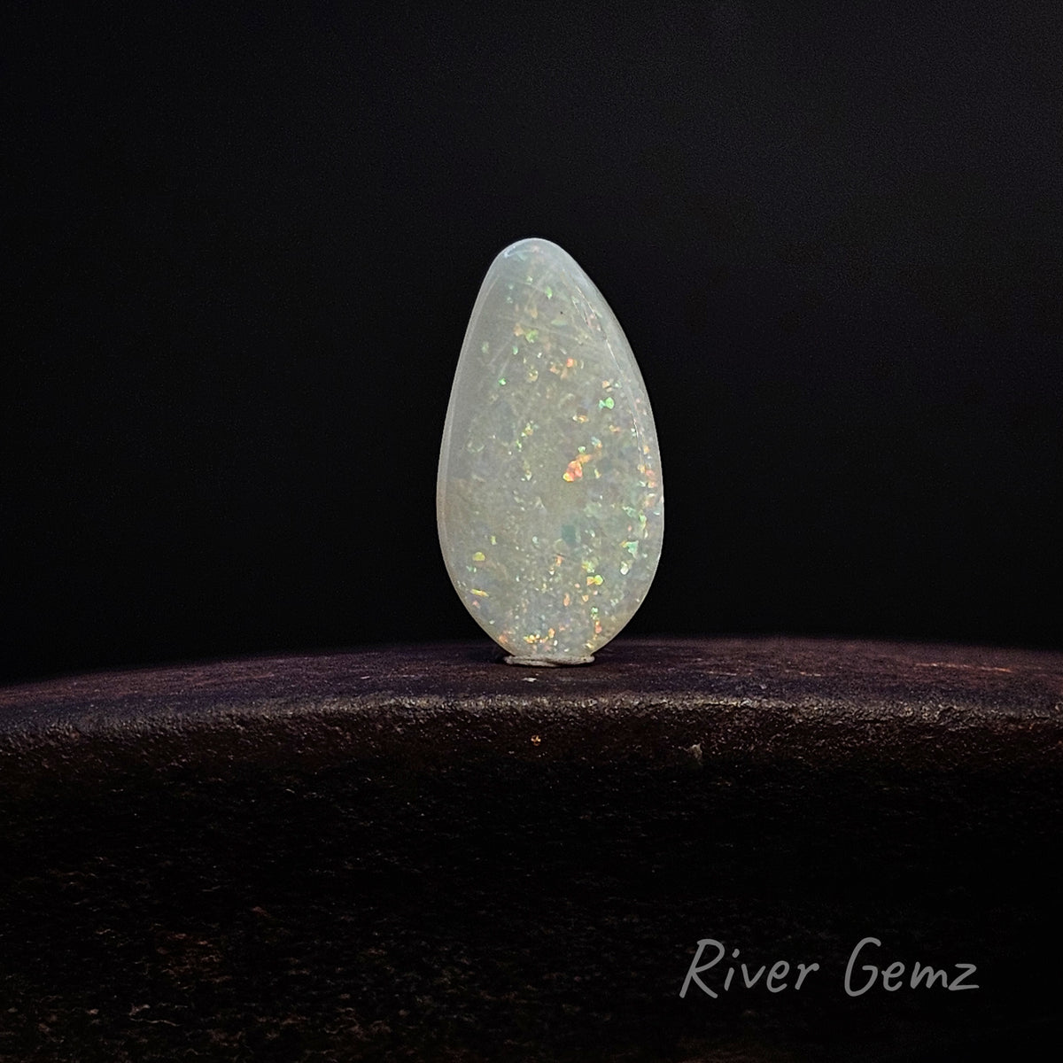 A teardrop-shaped white opal gemstone with iridescent flashes, displayed on a dark wooden surface against a black background. The 'River Gemz' brand logo is visible in the bottom right corner.