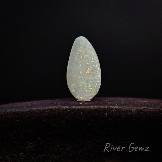A teardrop-shaped white opal gemstone with iridescent flashes, displayed on a dark wooden surface against a black background. The 'River Gemz' brand logo is visible in the bottom right corner.