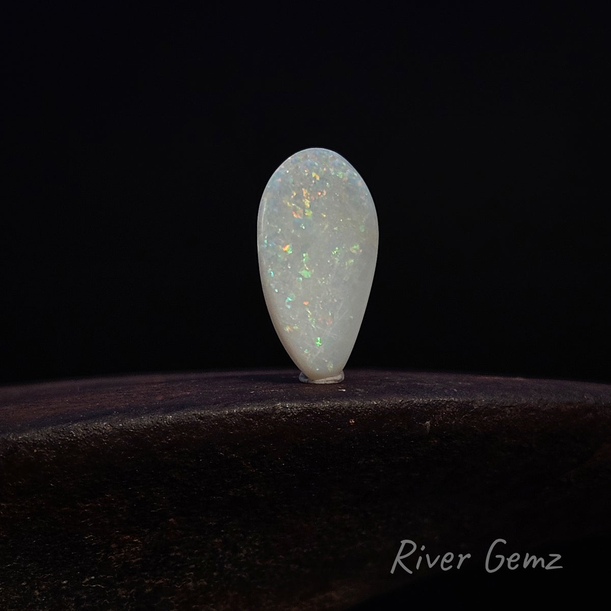 A teardrop-shaped white opal gemstone with iridescent flashes, displayed on a dark wooden surface against a black background. The brand name 'River Gemz' is visible in the bottom right corner.