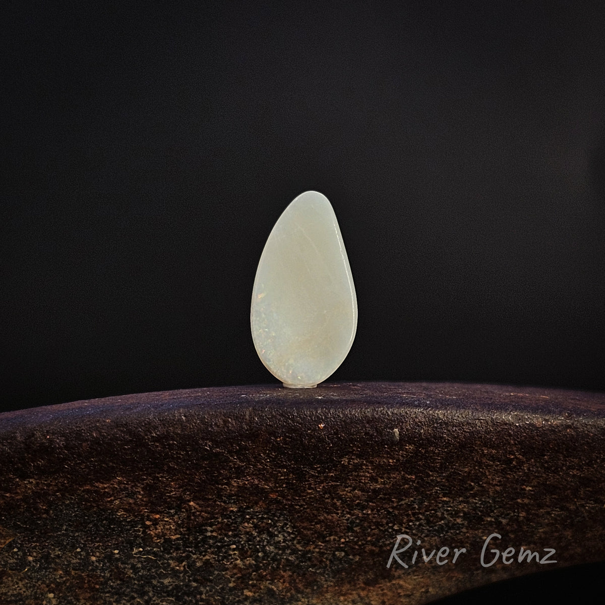 Rear view of a polished, teardrop-shaped opal to show light base tone of the white opal standing upright on a dark, textured wooden surface against a solid black background. The brand name 'River Gemz' is visible as a watermark in the bottom right corner.