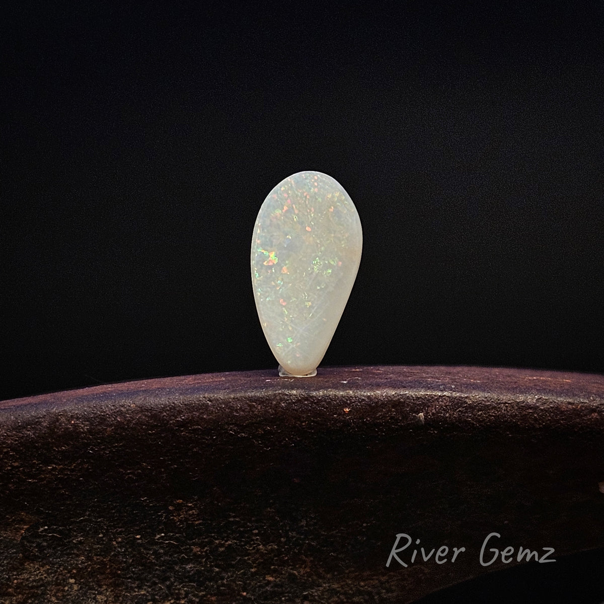 A teardrop-shaped white opal gemstone with iridescent flashes, displayed on a dark textured surface against a black background. The 'River Gemz' brand logo is visible in the bottom right corner.