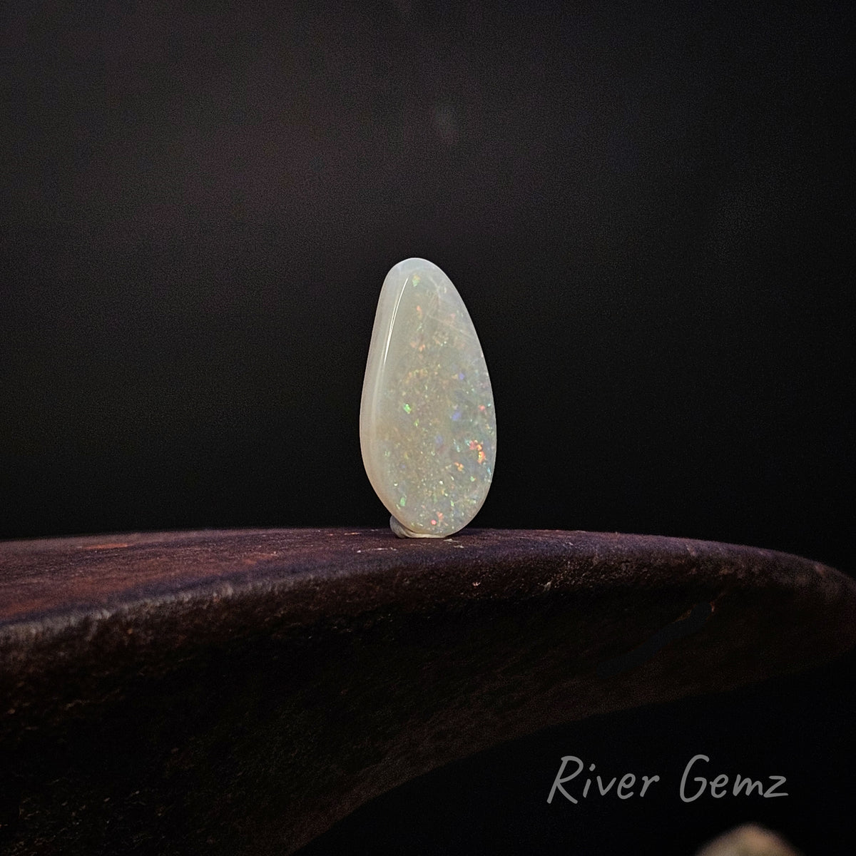 A teardrop-shaped cabochon gemstone with a milky white body and vibrant internal play-of-color flashes, standing upright on a dark wooden surface against a dark background. The 'River Gemz' brand logo is visible in the bottom right corner.
