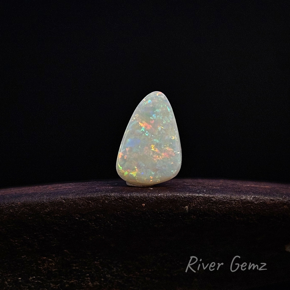A teardrop-shaped white opal gemstone with vibrant multicolor flashes, displayed on a dark wooden surface against a solid black background. The brand name 'River Gemz' is visible in the bottom right corner.