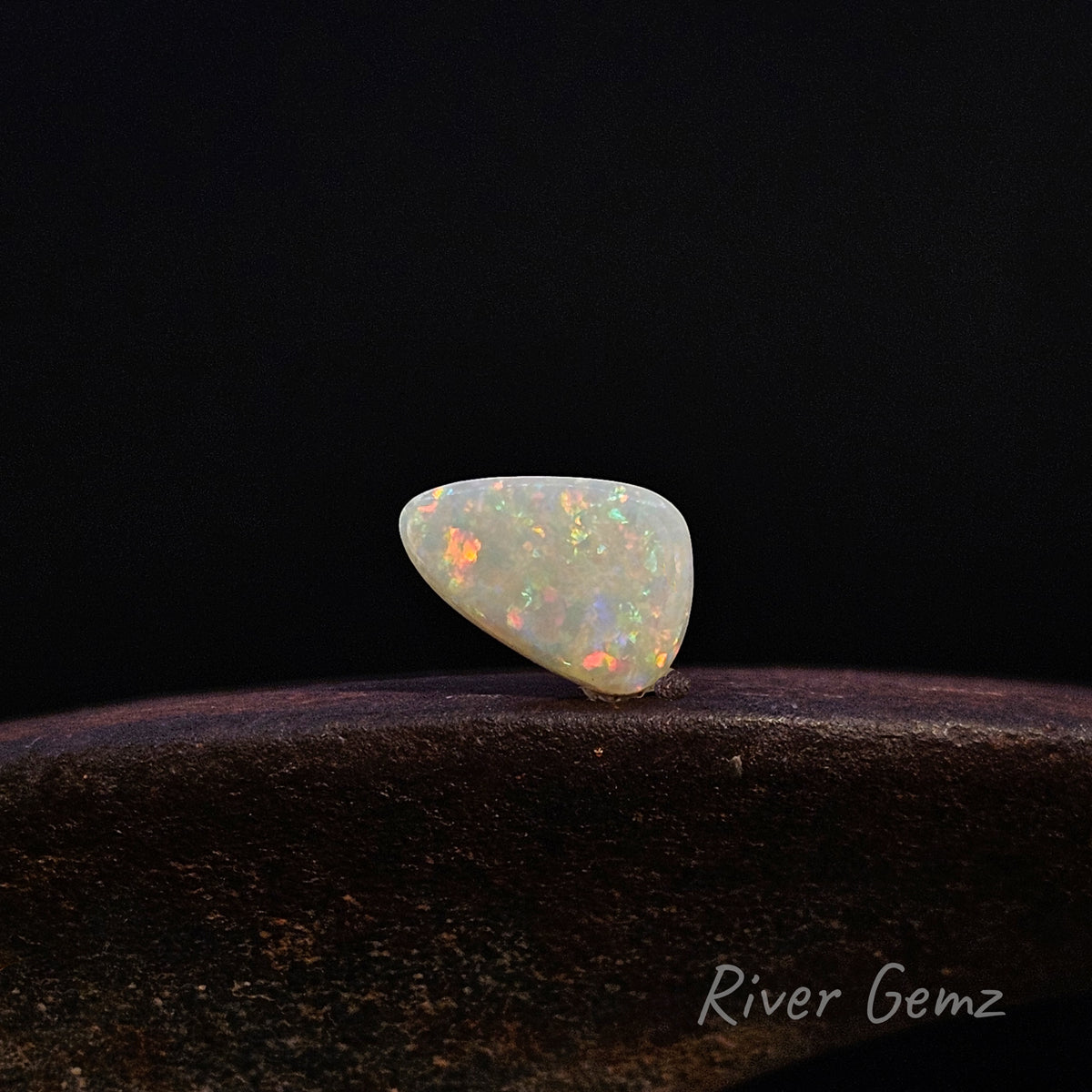 Free-form cut multi-coloured white opal resting on a weathered shoe anvil with 'River Gemz' watermark in the bottom right hand corner.