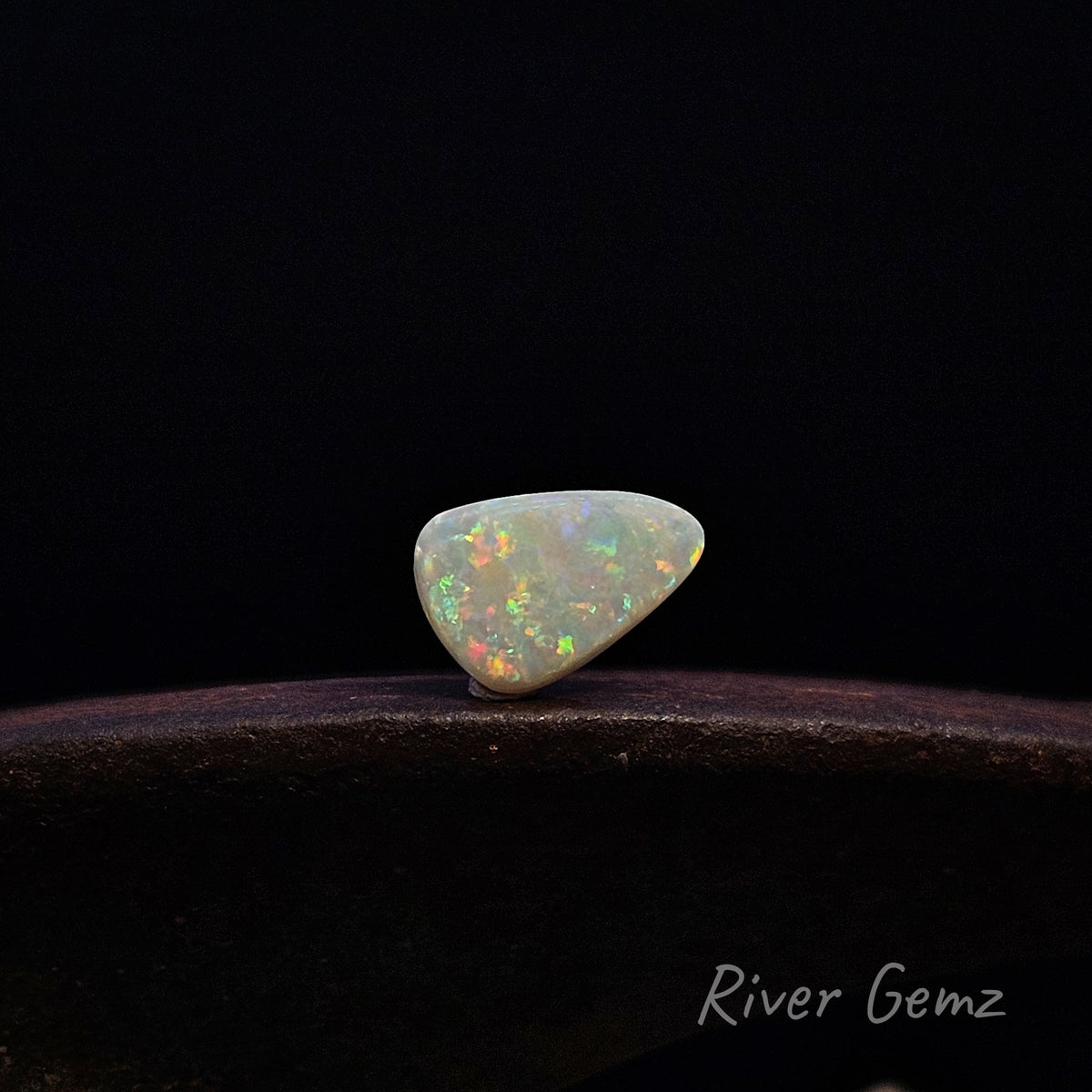 A small teardrop-shaped white opal gemstone with vibrant multicolor flashes, resting on a dark wooden surface against a solid black background. The brand name 'River Gemz' is visible in the bottom right corner.