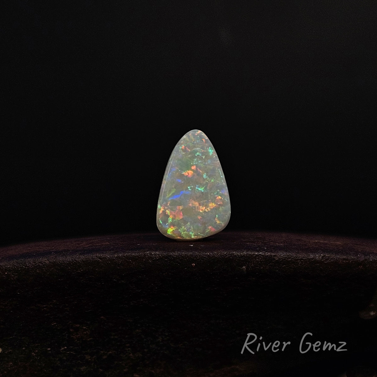 A teardrop-shaped white opal gemstone with vibrant multicolor flashes, displayed on a dark wooden surface against a black background. The 'River Gemz' brand logo is visible in the bottom right corner.