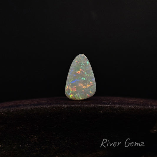 A teardrop-shaped white opal gemstone with vibrant multicolor flashes, displayed on a dark wooden surface against a black background. The 'River Gemz' brand logo is visible in the bottom right corner.