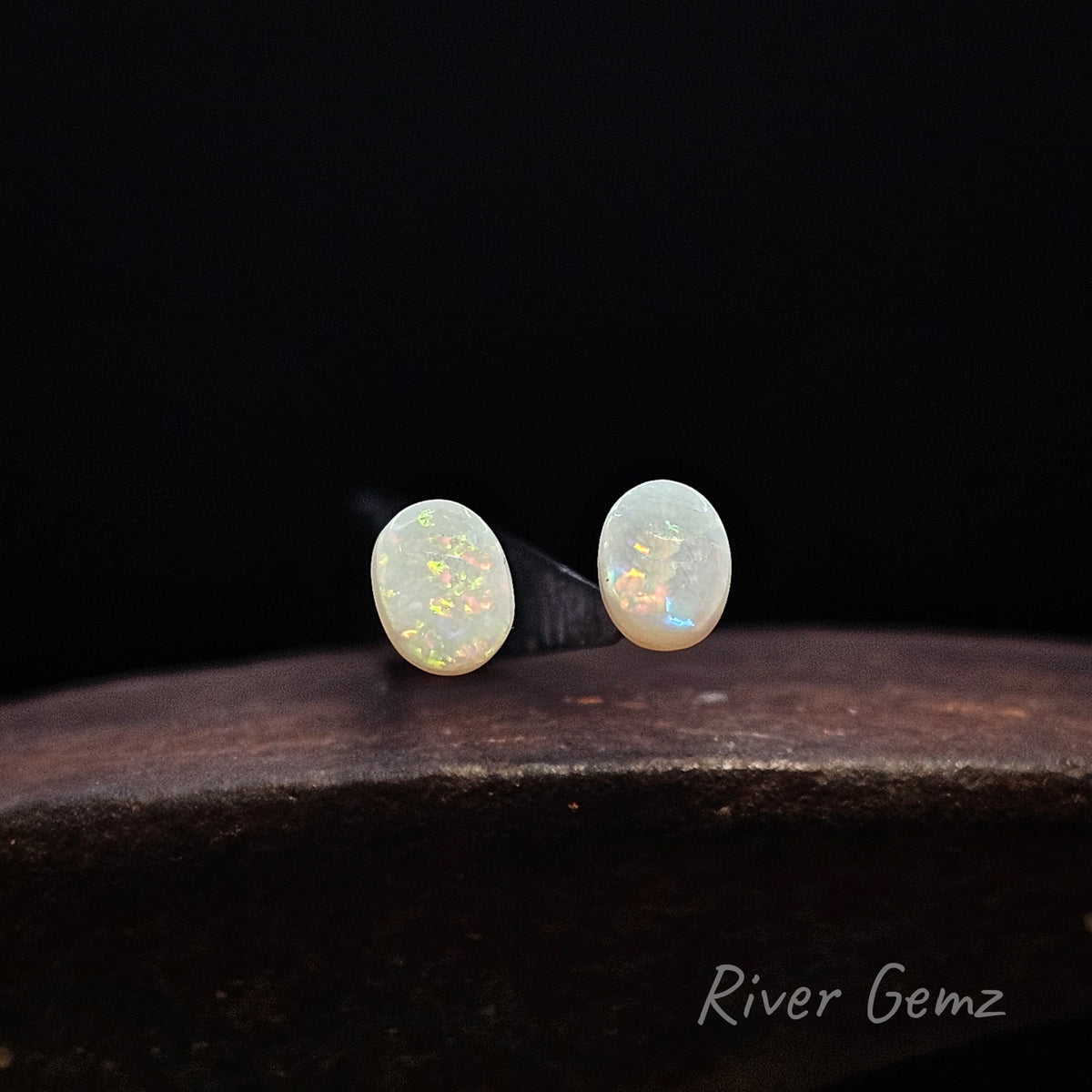 Pair of unset oval shaped multi-coloured white opals against a black background and 'River Gemz' watermark in the bottom right hand corner.