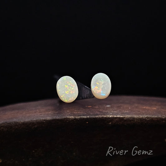Pair of unset oval shaped multi-coloured white opals against a black background and 'River Gemz' watermark in the bottom right hand corner.