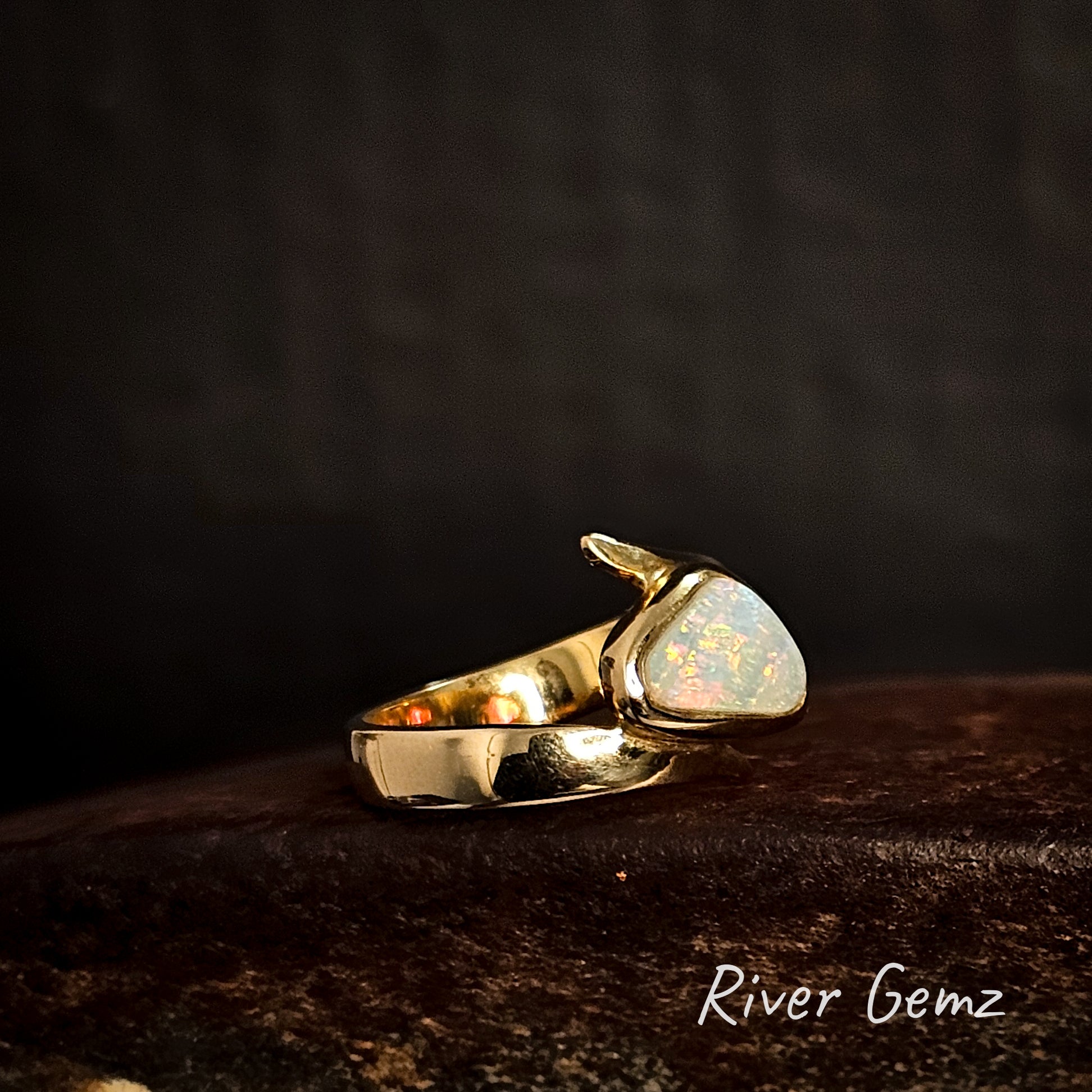 Gold ring with an opal stone on a dark background, featuring the brand 'River Gemz'.