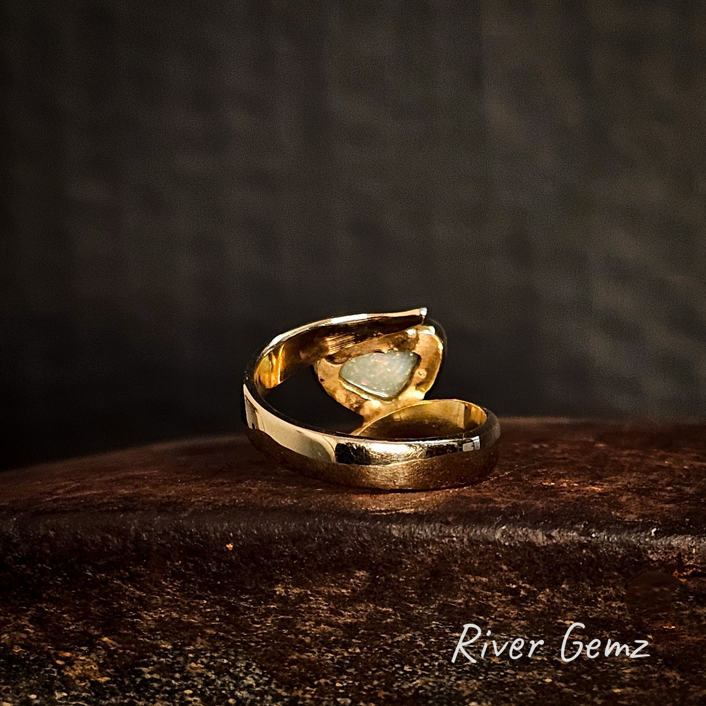 Back of gold ring showing the light base tone of the included white opal on a cast iron surface, branded 'River Gemz'.