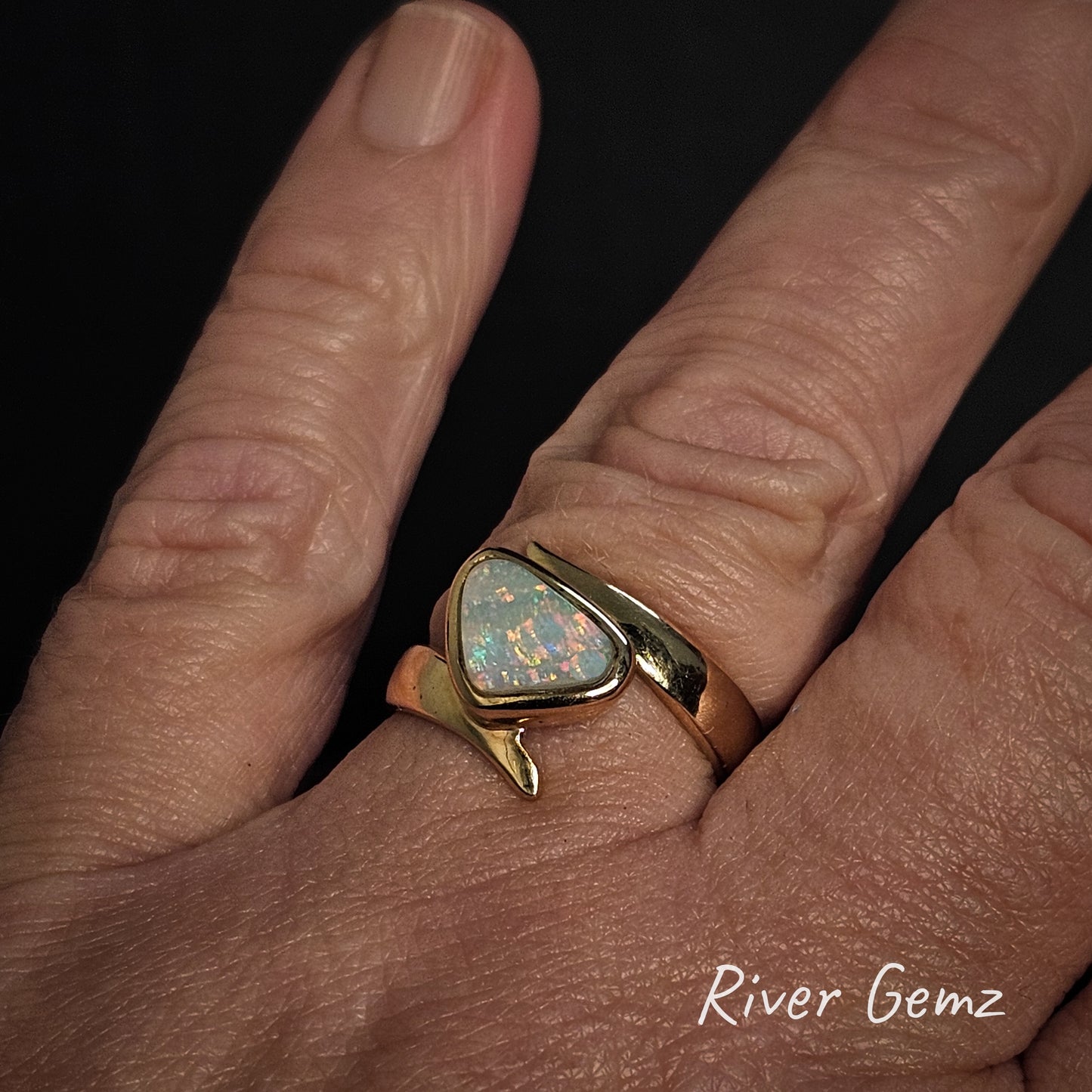 Gold ring with an opal stone on the ring finger of a hand, branded 'River Gemz'.
