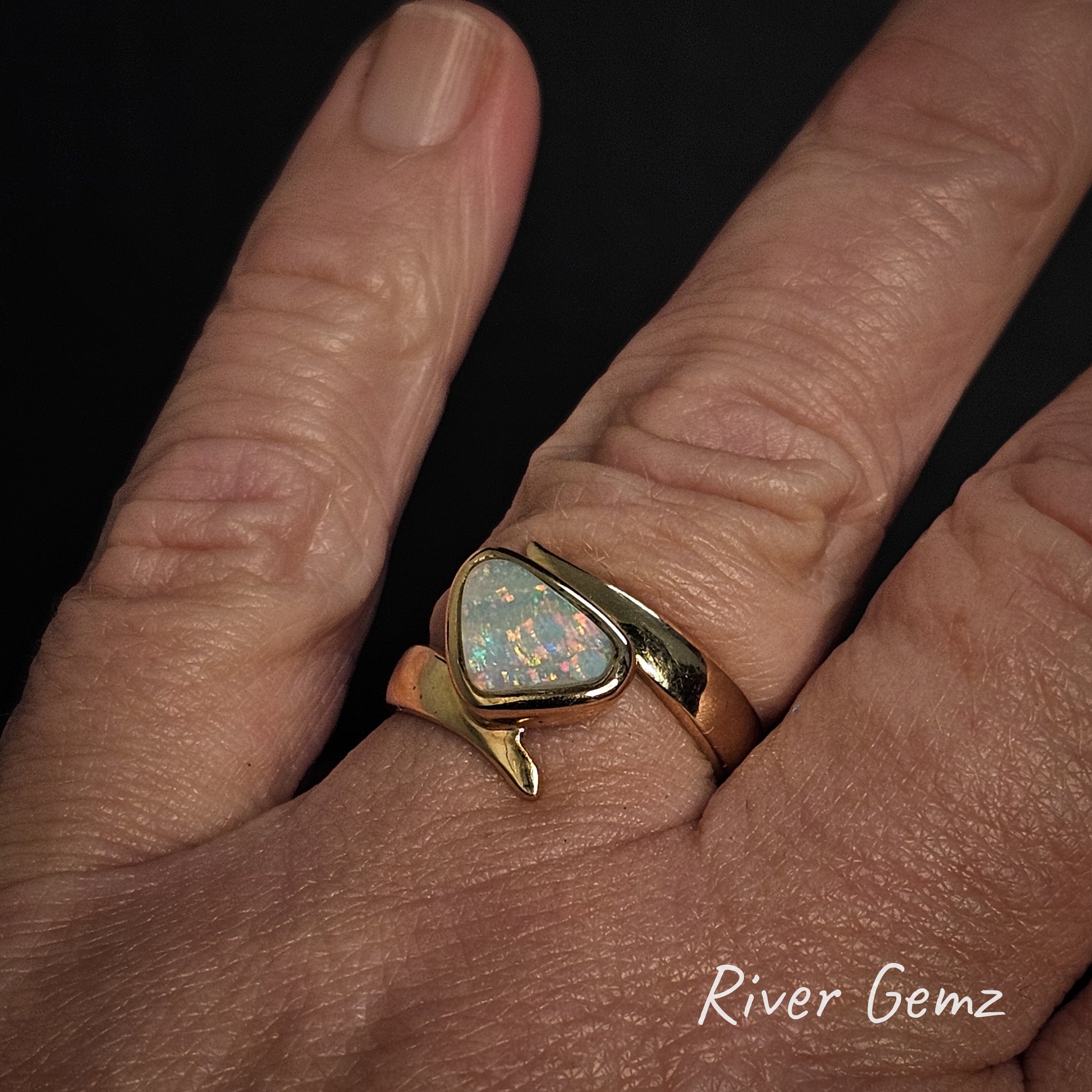 Gold ring with an opal stone on the ring finger of a hand, branded 'River Gemz'.